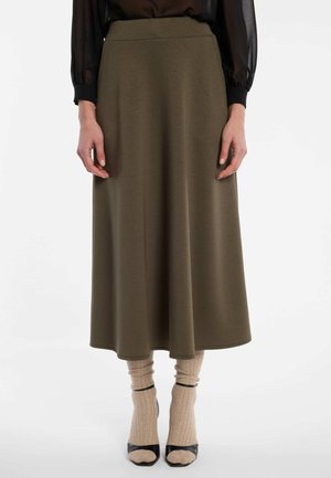 Frau trägt einen olivgrünen Midirock, beige Socken, schwarze offene High Heels und eine schwarze transparente Langarmbluse.