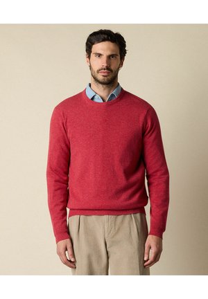 Roter Strickpullover mit Rundhalsausschnitt und gerippten Bündchen, kombiniert mit einem hellblauen Hemd mit Kragen. Dazu beige Cordhosen.