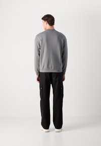 En mann som står med ryggen til, iført en grå langermet genser, svarte cargo-bukser og hvite sneakers mot en ensfarget bakgrunn.