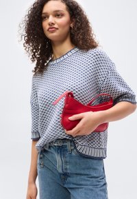 Νέα γυναίκα με κατσαρά μαλλιά, που φοράει μπλε πουλόβερ με σχέδιο και κοντά μανίκια και τζιν, κρατώντας μια μικρή κόκκινη τσάντα στη μέση.