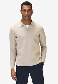 Beige kofta med kvartdragkedja, platt krage och ribbad struktur. Långa ärmar, bärs över en vit skjorta, med en figurnära design.