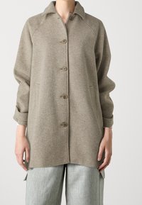 Persoon draagt een beige wollen jas met knopen, voorzakken en lange mouwen, gecombineerd met lichtgrijze broek met zijzakken.