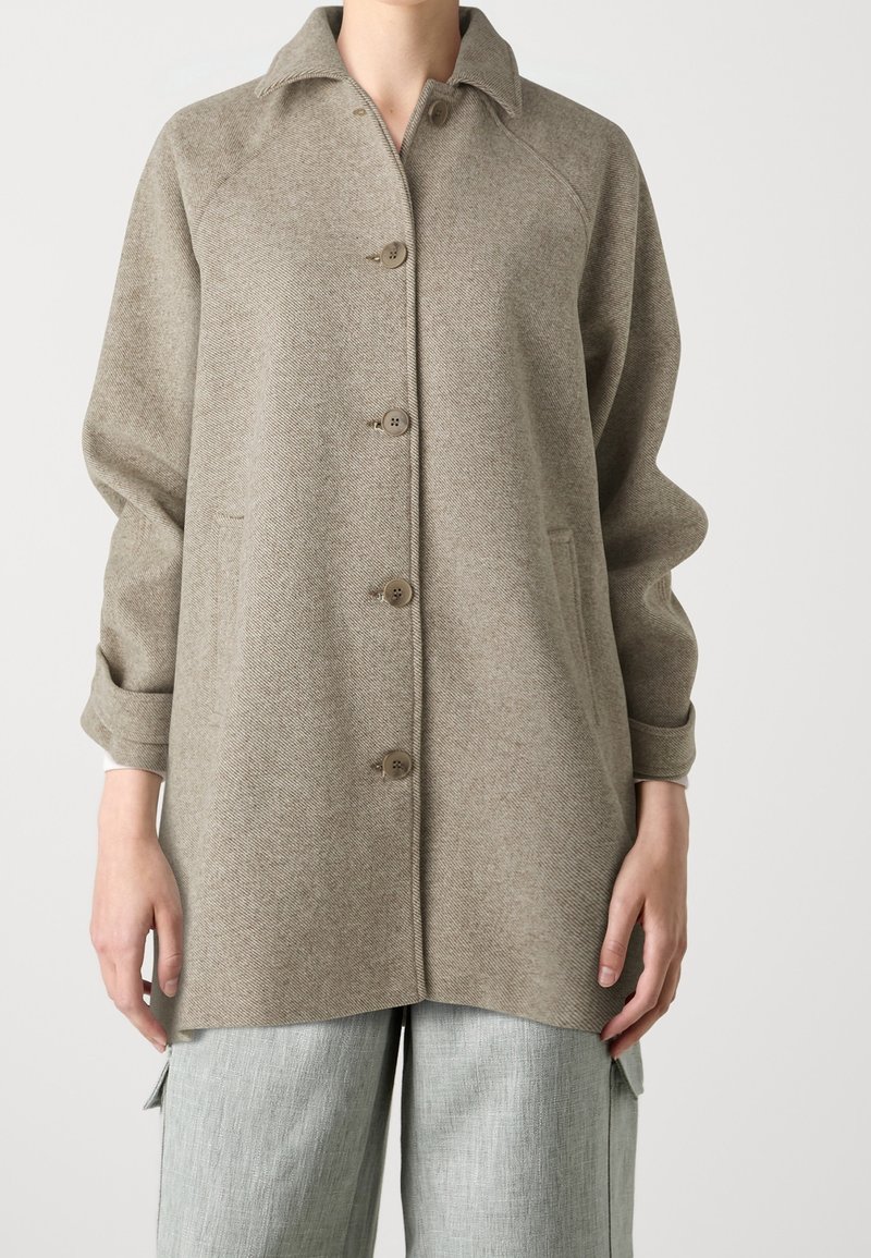 Persoon draagt een beige wollen jas met knopen, voorzakken en lange mouwen, gecombineerd met lichtgrijze broek met zijzakken.