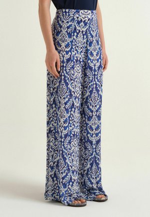 Donna che indossa pantaloni a gamba larga con un complesso motivo floreale blu e bianco, abbinati a top nero e sandali open toe.