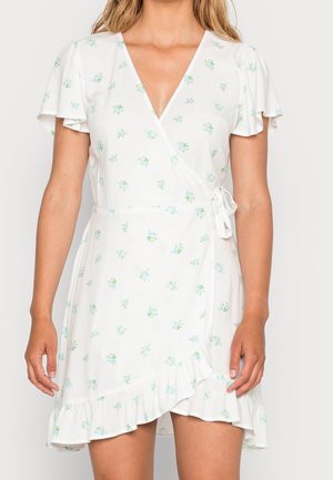 Femme portant une robe portefeuille blanche avec de petits imprimés floraux bleus, des manches courtes volantées et un ourlet à volants, nouée à la taille sur le côté droit.