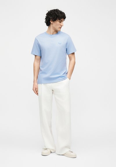 Moški s kodrastimi temnimi lasmi, oblečen v svetlo modro majico s kratkimi rokavi, bele široke hlače in bele superge, stoji z eno roko v žepu.