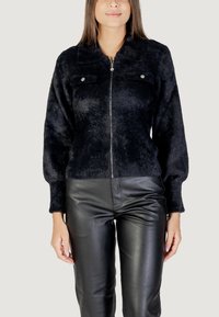 Veste zippée en velours noir avec de longues manches bouffantes, dotée de deux poches poitrine et de quincaillerie argentée, associée à un pantalon en cuir noir ajusté.