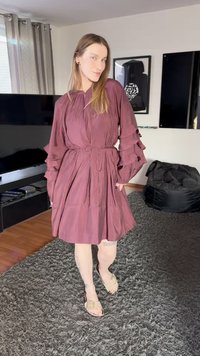 Fialové šaty s volánkovými rukávy, stahovaným pasem a rozšířenou sukní. Nošeny s lehkými sandály na texturovaném koberci v obývacím pokoji.