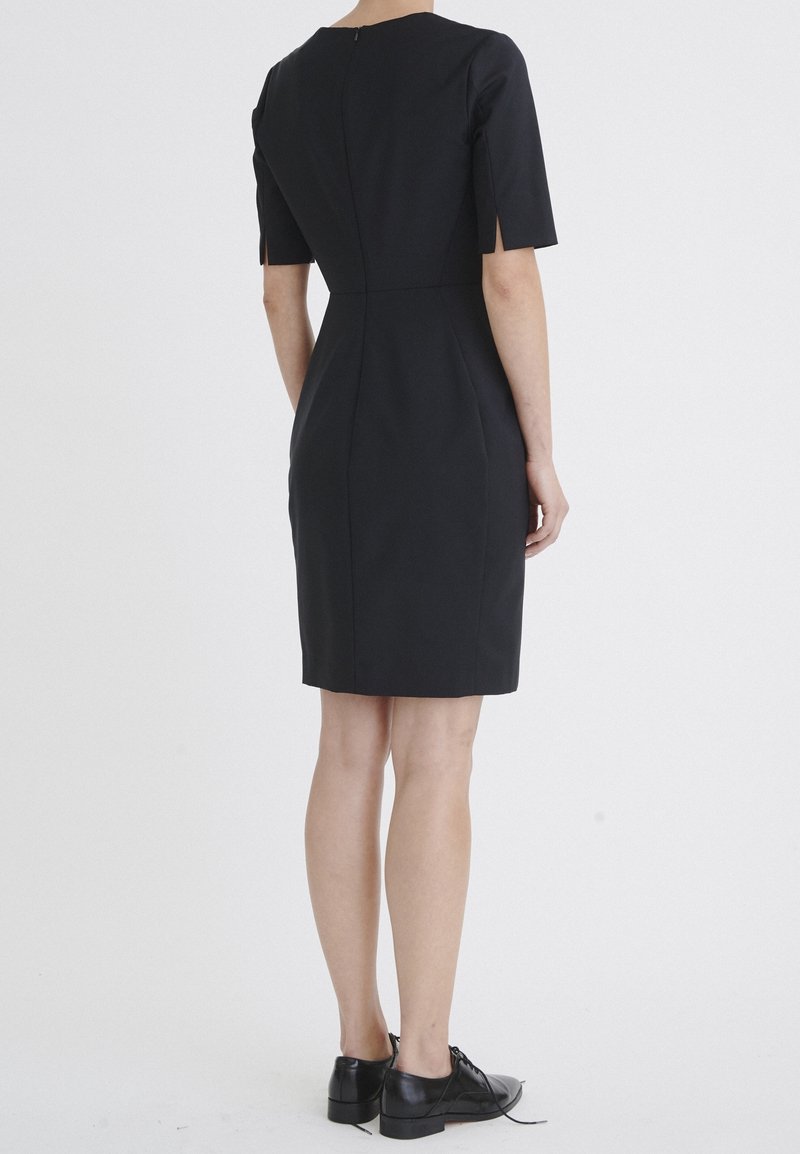 Svart figurnära klänning med korta ärmar, dragkedja bak och skräddarsydda sömmar. Bärs med svarta skor och visar en knälång design.