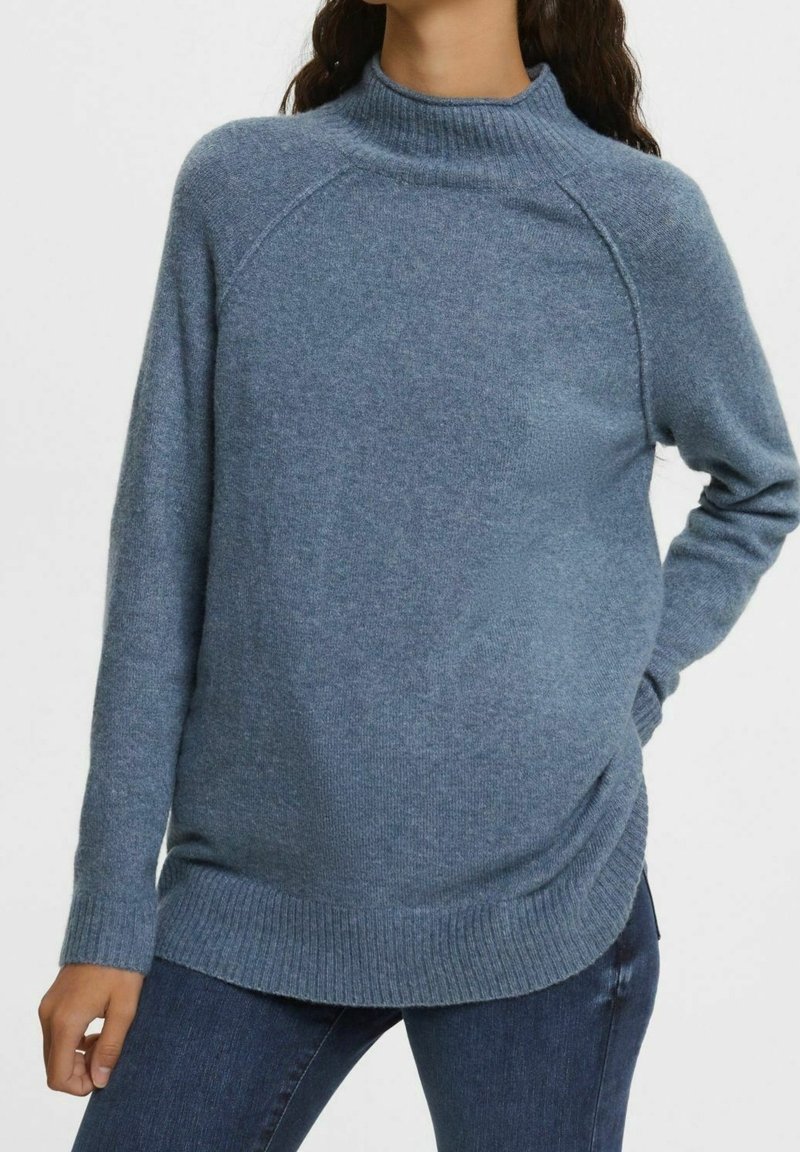 Blå stickad tröja med hög krage, ribbade mudd- och nederkanter, med en avslappnad passform och subtil struktur. Kombinerad med mörka jeans.