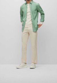 Ljusgrön skjorta över en krämfärgad tröja, kombinerad med beige byxor och vita sneakers, visad mot en grå bakgrund.