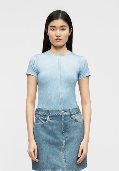 Γυναίκα με μακριά σκούρα μαλλιά, φοράει ανοιχτό μπλε εφαρμοστό κοντομάνικο μπλουζάκι και σκουρόχρωμη τζιν φούστα, μπροστά από απλό γκρι φόντο.