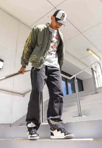 Chaqueta bomber oliva con bordado amarillo, camiseta gráfica blanca, jeans negros y zapatillas en blanco y negro en una escalera.