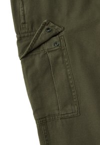 Nahaufnahme eines olivgrünen Stoffes mit einer rechteckigen Tasche, die eine Klappe besitzt, die mit zwei schwarzen Druckknöpfen gesichert ist.