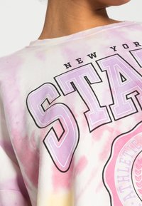 Camiseta de algodón con un patrón de tie-dye en rosa y amarillo. Texto en negrita "STATE" en púrpura con un contorno negro, detalle del logo redondo debajo.