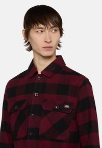 Camisa de franela a cuadros rojos y negros con cuello, dos bolsillos en el pecho y cierre de botones. Fabricada en tela suave y texturizada.