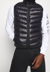 Schwarze Steppweste mit gesteppter Optik, glänzender Textur, durchgehendem Reißverschluss und hohem Kragen, getragen über einem weißen Langarmshirt.