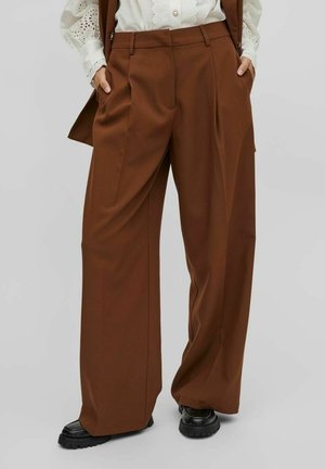 Vrouw draagt hooggesneden brede bruine broek met zijzakken, gecombineerd met een witte blouse met oogjes en zwarte chunky schoenen.