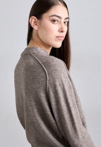 Nuori nainen, jolla on pitkät ruskeat hiukset, pukeutuneena harmaaseen neulepaitaan, katsoo olkansa yli vaaleanharmaata taustaa vasten.