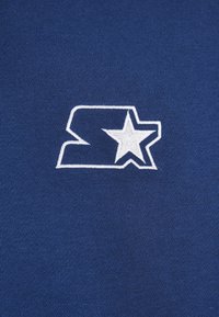 Námořnická modrá látka má bílé logo se znakem "S" a hvězdou. Textura je hladká s mírným pružením.