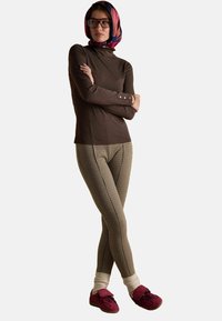 Nais, kellel on seljas värvikas pearä the, pruun pikka varrukatop, mustrilised retuusid, kreemjad sokid ja punased tossud, seisab käed ristis.