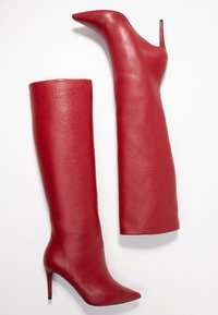 Bottes en cuir rouge montantes, avec une texture lisse, un bout pointu et un talon aiguille. Une botte a un design à côté ouvert.