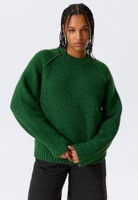Jong persoon met gevlochten haar, gekleed in een groene oversized wollen trui en zwarte broek, staande tegen een witte achtergrond.