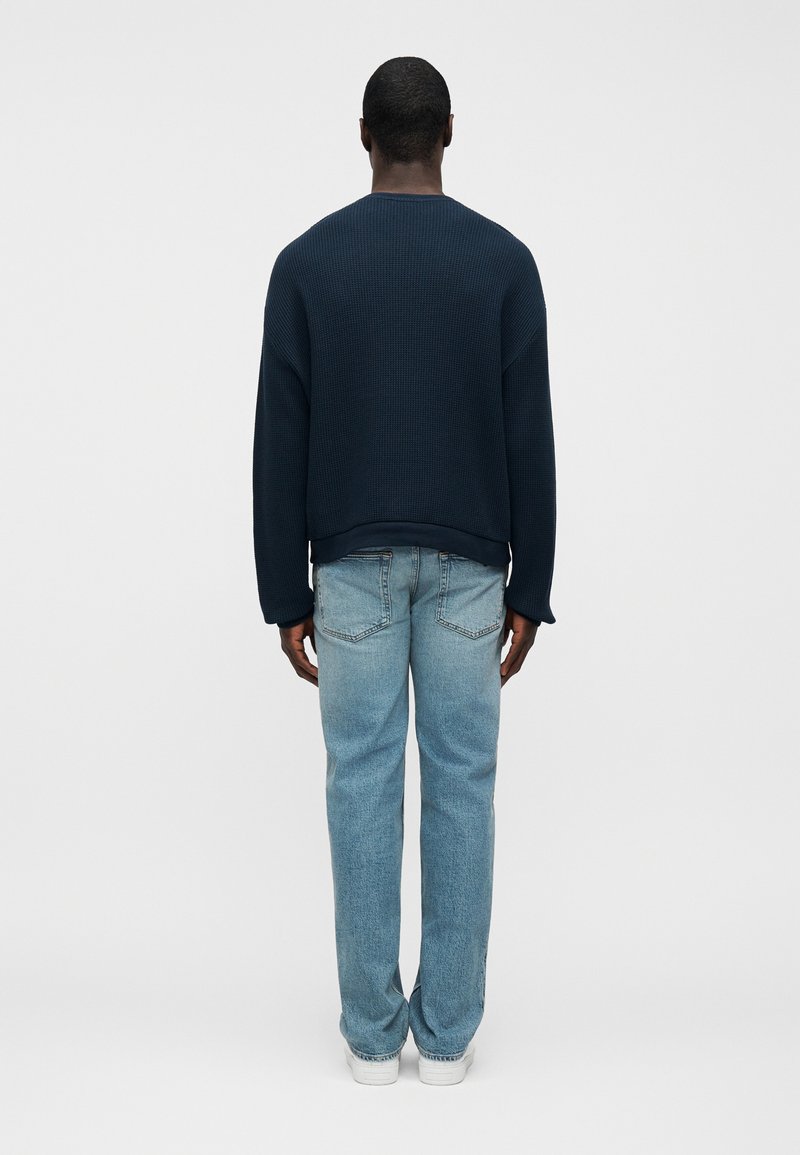 Pull en maille texturée bleu marine avec un col rond et des épaules tombantes, associé à un jean droit bleu clair et des baskets blanches.