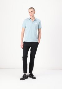 Světle modrá polo košile s krátkým rukávem a límečkem, zkombinovaná s černými slim-fit kalhotami a černými botami, stojící na bílém pozadí.