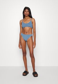 Biquíni azul elástico com um top cropped e parte de baixo de cintura alta. Textura suave, design minimalista, combinado com sandálias deslizantes pretas.