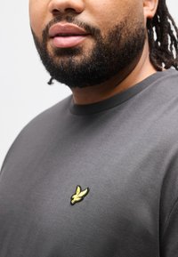 Άνδρας με γένια που φοράει σκούρο γκρι πουκάμισο με μικρό κίτρινο λογότυπο πουλιού στο στήθος, εμφανιζόμενος από το πηγούνι έως το άνω μέρος του κορμού.