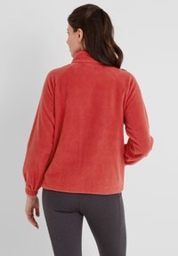Coral fleece jacket with long sleeves, high collar, and elastic cuffs. Smooth texture and relaxed fit, paired with dark gray leggings.