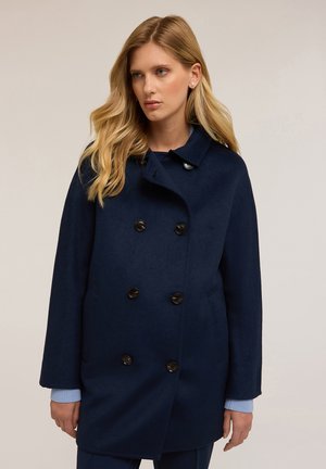 Marineblauer Wollpeacoat mit doppelt geknöpftem Verschluss, seitlichen Taschen und einem wattierten Reverskragen. Hellblaue Strickbündchen sind sichtbar.