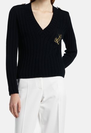Femme portant un pull noir à côtes en V avec un emblème doré sur la poche poitrine et un pantalon blanc taille haute, debout, un bras derrière le dos.