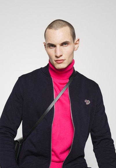 Tumši zils zip-up džemperis pār spilgti rozā augsto apkakli. Gluda auduma virsma ar nelielu izšuvumu uz džempera. Melns pārslēdzams siksna.