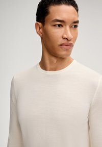 Maglia a maniche lunghe color crema in maglia con scollo rotondo, texture liscia e dettagli di cuciture sottili sulle maniche e sullo scollo.