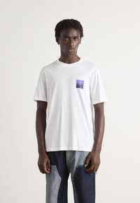 White short-sleeve t-shirt with a purple rectangular pocket featuring the logo "HUGO" in white, paired with blue denim jeans.