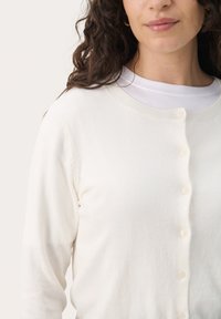 Vaalea kerma cardigan, joka on valmistettu pehmeästä kankaasta, pyöreällä kauluksella, pitkillä hihoilla ja viidellä napilla edessä. Sileä tekstuuri.