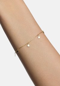 Złota bransoletka łańcuszkowa z małymi zawieszkami w kształcie gwiazdek, gładka w dotyku, delikatne ogniwa i zapięcie, noszona na jasnej karnacji.