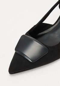 Zapato de ante negro con punta afilada que presenta un destacado acento de hardware brillante negro en el empeine y una elegante correa en el tobillo. Textura suave en general.