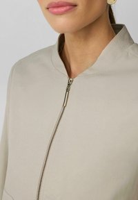 Veste beige à fermeture éclair avec col Mandarin, présentant une texture lisse et une fermeture centrale. Le tirette en métal ajoute une touche subtile.