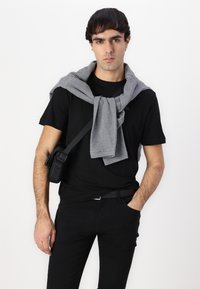 Fekete póló, szürke pulcsi a nyakban, fekete nadrág, és egy kis fekete válltáska. A modell az egyik kezét a csípőjén tartja.