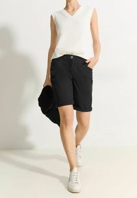 Schwarze Shorts mit aufgekrempelten Bündchen, kombiniert mit einem strukturierten weißen ärmellosen Oberteil. Modell trägt weiße Sneaker und hält eine schwarze Jacke.