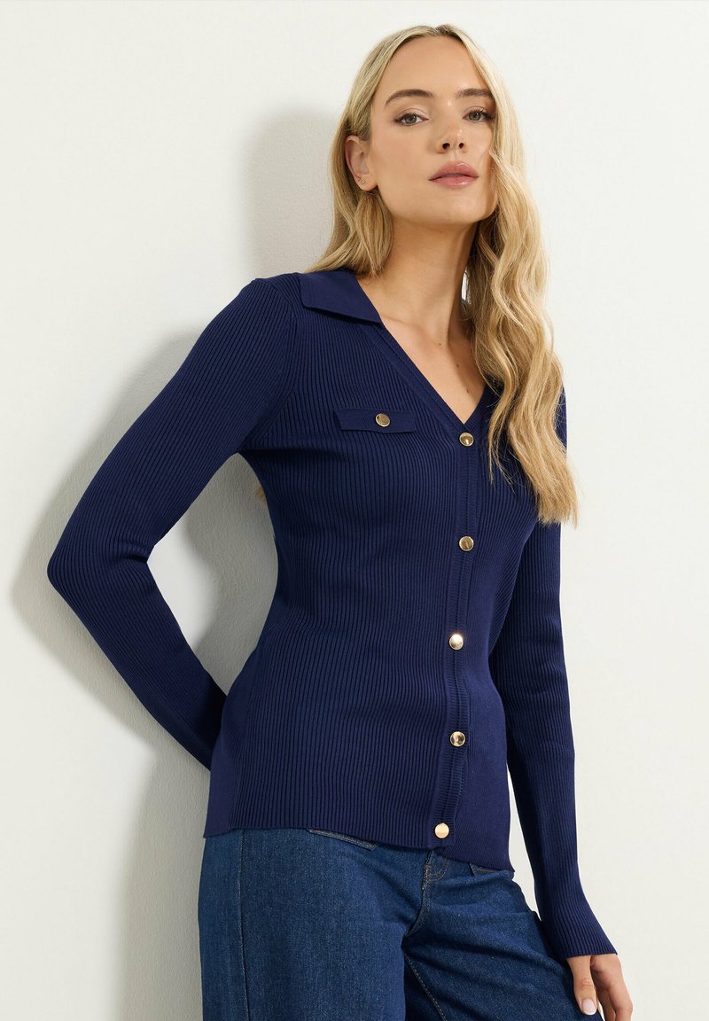 Mörkblå ribbad stickad cardigan med V-ringning, knappfram och guldfärgade knappar. Har krage och sidofickor. Långa ärmar.