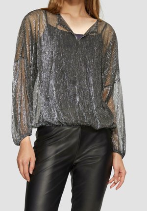 Vrouw met een zilverkleurige metalen doorzichtige blouse met lange mouwen en kraag en zwarte leren broek, afgebeeld van nek tot halverwege de dij.