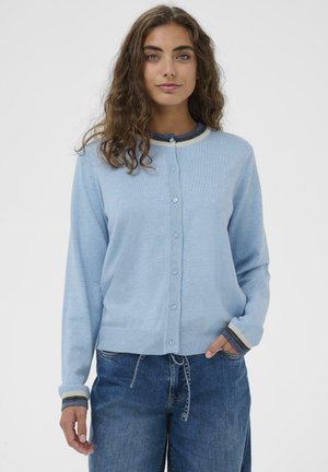 Jasnoniebieski kardigan z miękkiego materiału, z okrągłym dekoltem, zapięciem na guziki i prążkowanymi mankietami w paski. Noszony na ciemnych dżinsach.