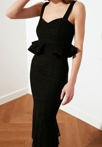 Abito in pizzo nero con silhouette aderente, dettaglio a peplum in vita e spalline larghe. Presenta un tessuto testurizzato con motivi floreali.