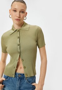 Top corto a maniche corte color verde oliva a coste con colletto, dotato di chiusura a bottone frontale e una linea aderente.