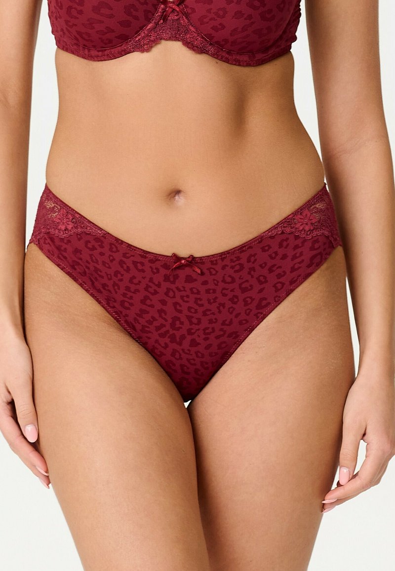 Femme portant un soutien-gorge et une culotte assortis imprimé léopard rouge foncé avec bordure en dentelle et petit nœud à l’avant de la culotte.