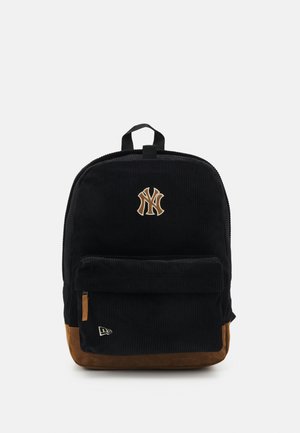 Sac à dos en velours côtelé noir avec un fond beige et une poche zippée à l'avant. Il est doté d'un logo brodé et d'une poignée en haut pour le transport.